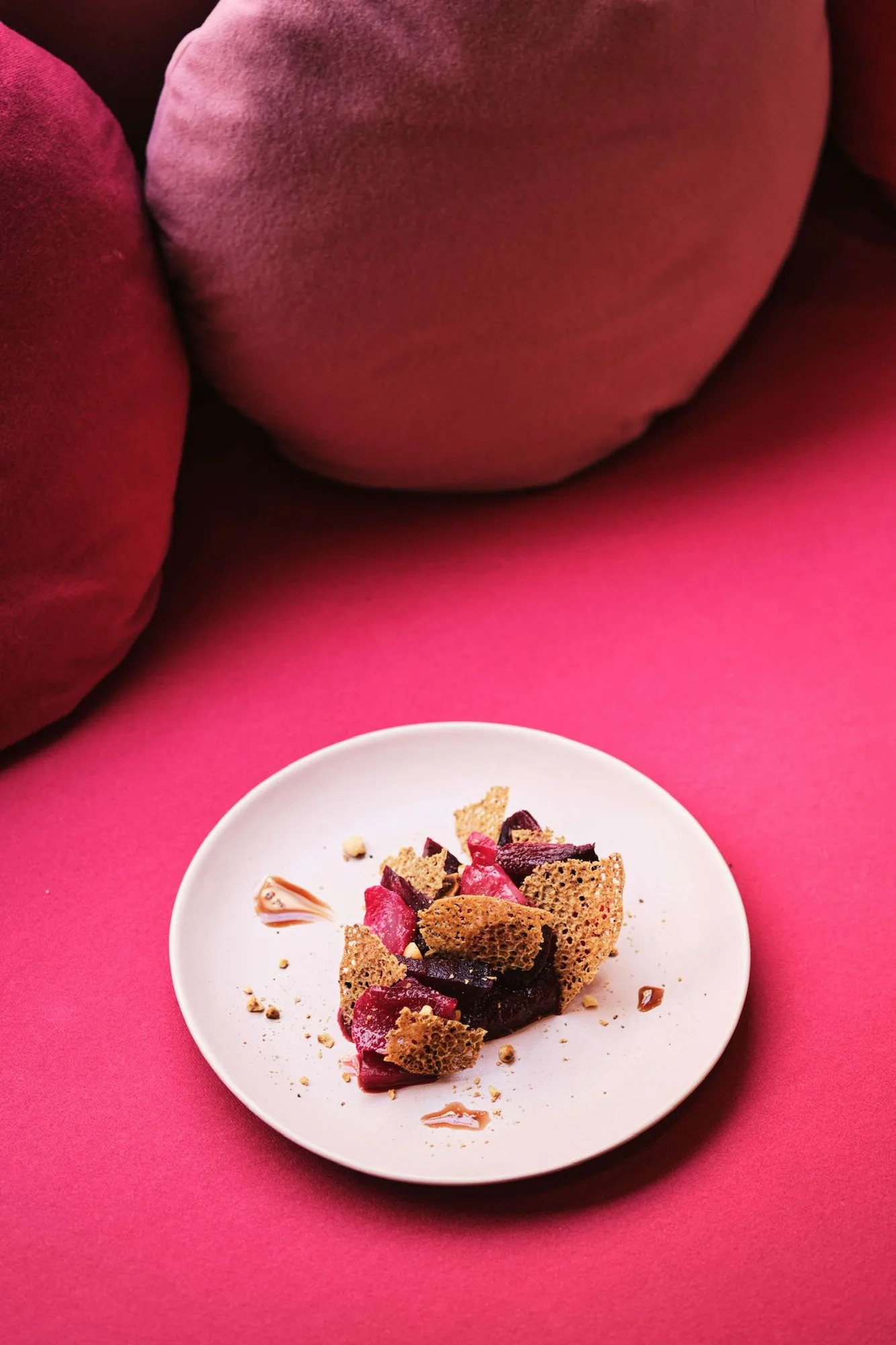 Assiette bistronomique : morceaux de betterave rôtie et tuiles croustillantes dorées sur fond fuchsia, coussins velours en arrière-plan.
