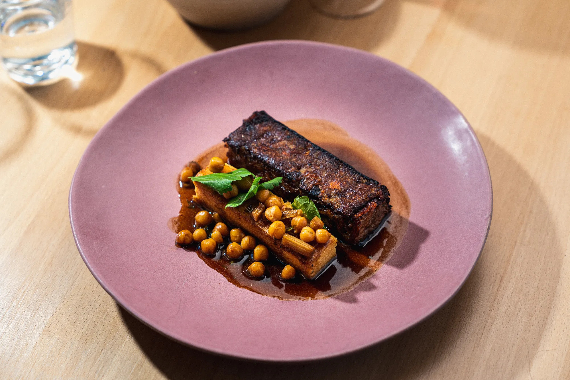 Assiette bistronomique rose : pièce de viande caramélisée, pois chiches rôtis et jus corsé, sur table en bois clair.