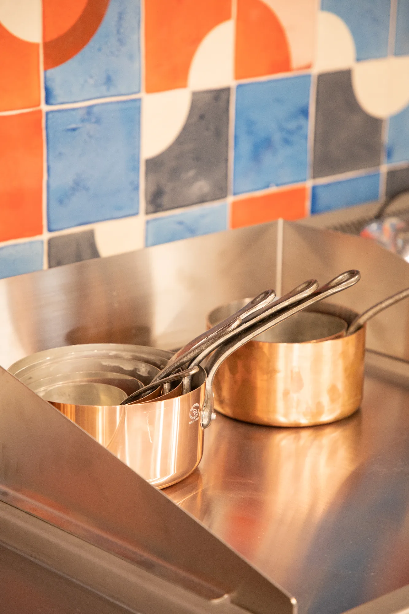 Casseroles en cuivre alignées sur un piano inox, devant un carrelage géométrique orange et bleu de la cuisine du restaurant HABILE.
