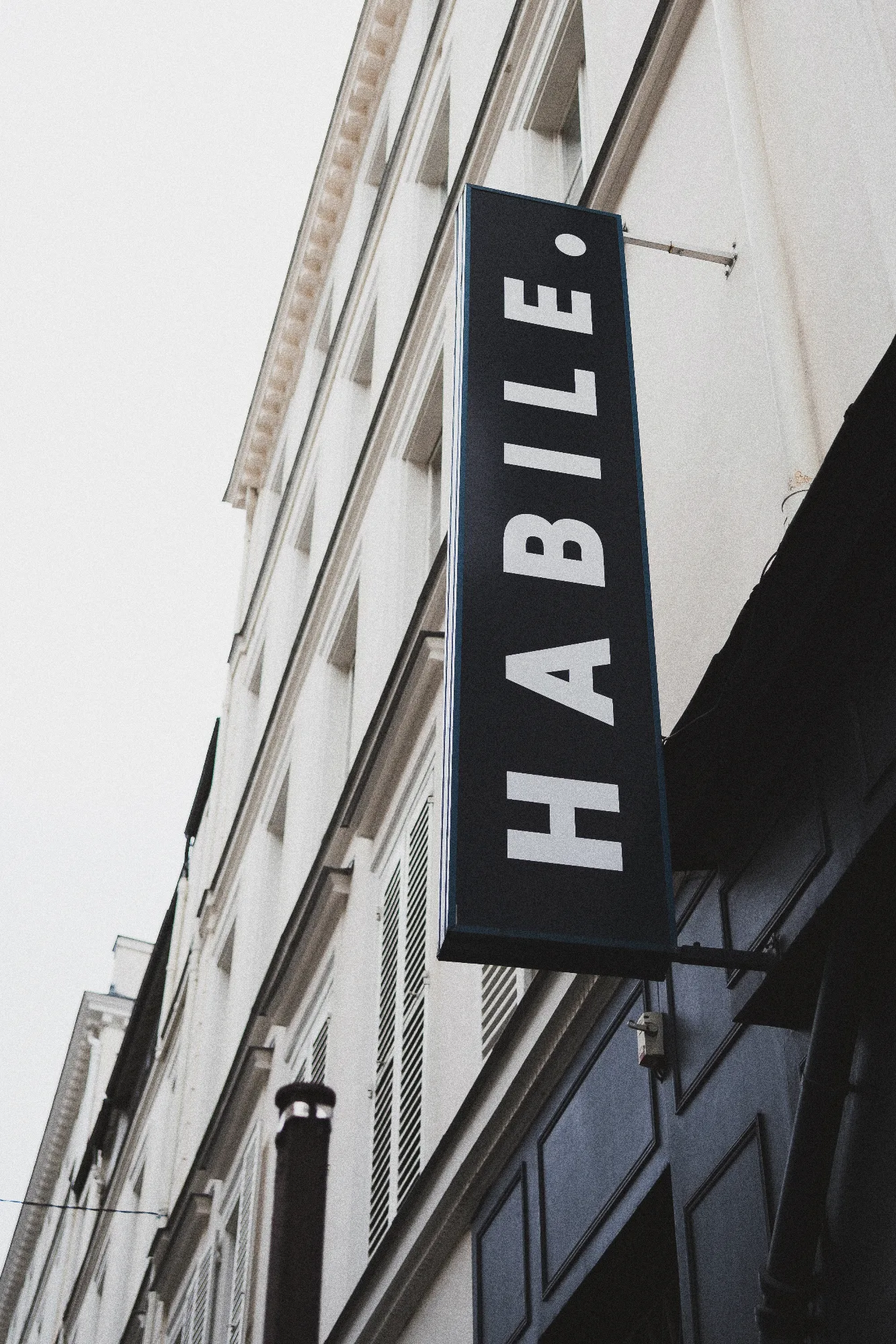 Enseigne verticale noire « HABILE. » fixée en façade d'un immeuble haussmannien, rue de Lancry, Paris 10.