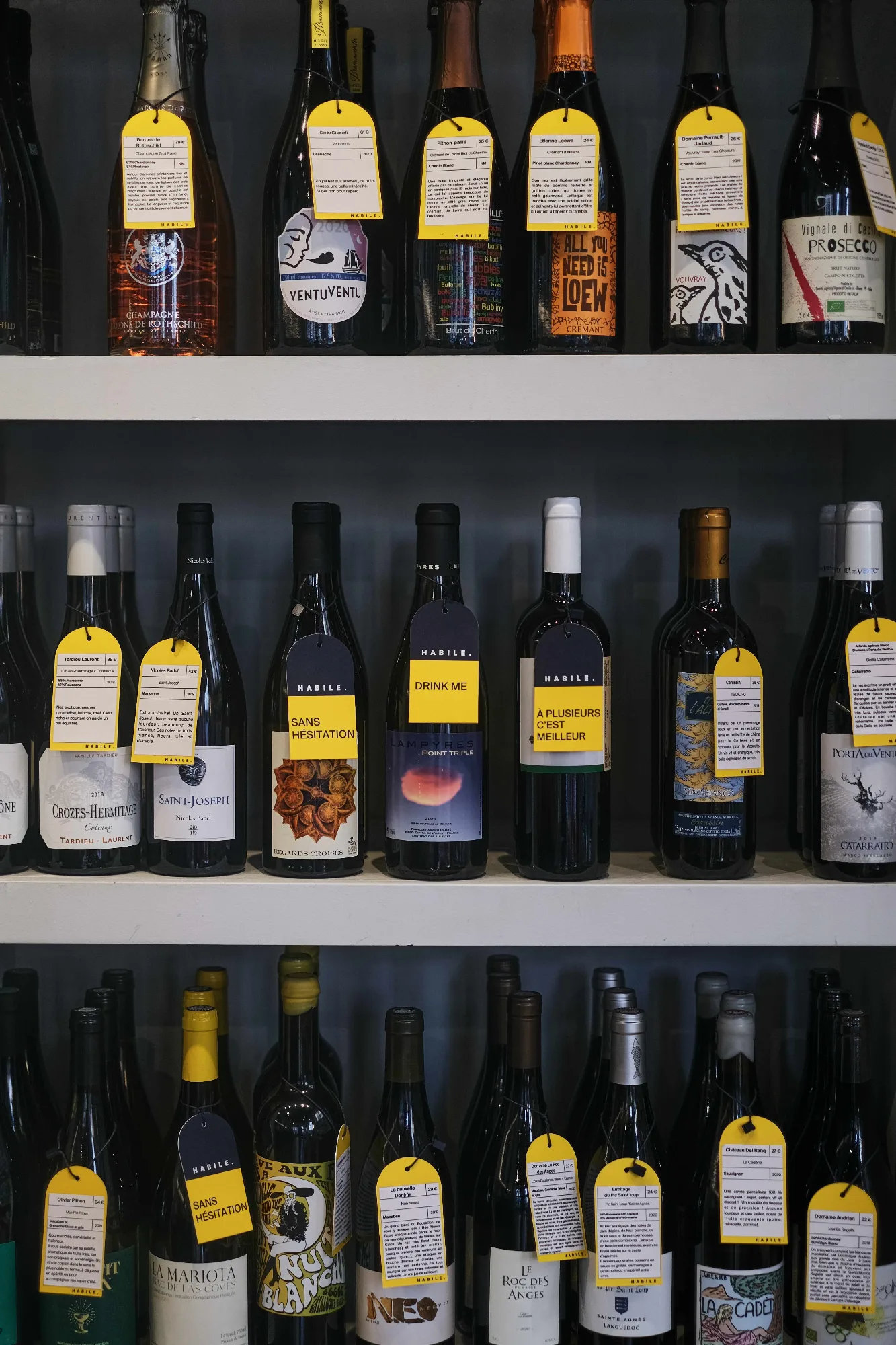 Étagères de la cave d'HABILE. garnies de bouteilles de vins vivants, chacune accompagnée d'une étiquette jaune manuscrite.