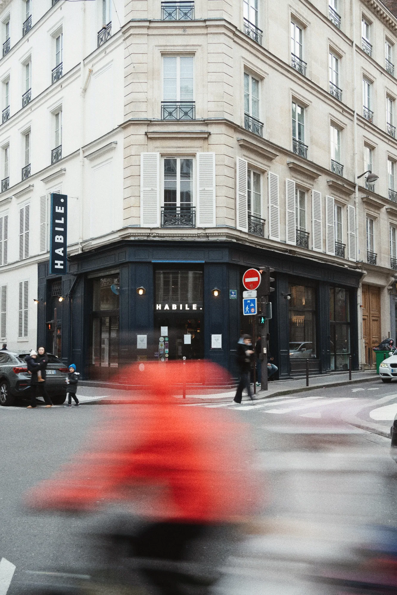 Façade d'HABILE. à l'angle d'un immeuble haussmannien du Paris 10, devanture bleu nuit animée par le flou d'un scooter rouge au premier plan.