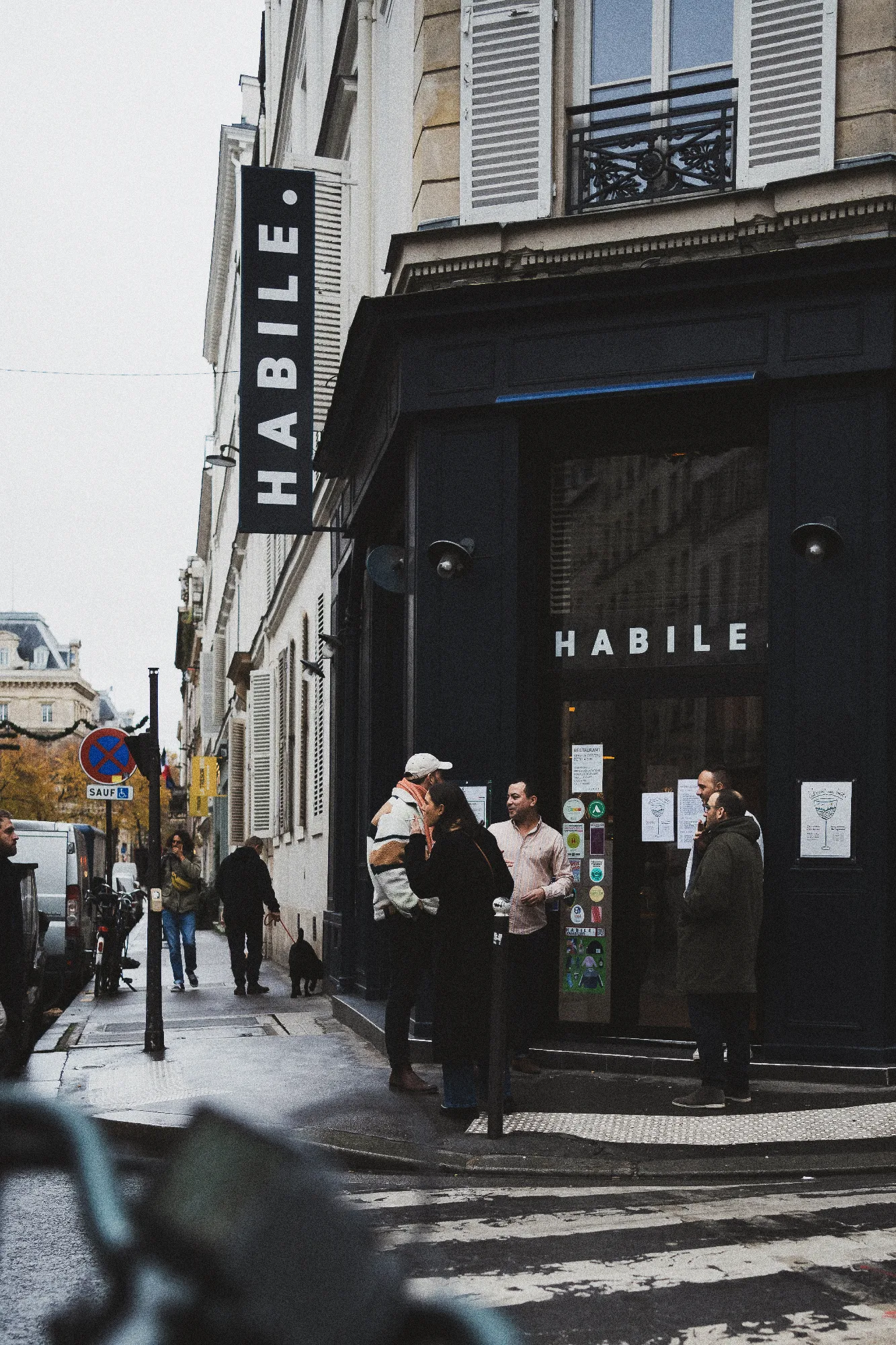 Façade sombre du restaurant HABILE. rue de Lancry, avec quelques passants attablés devant l'entrée, sous un ciel parisien couvert.