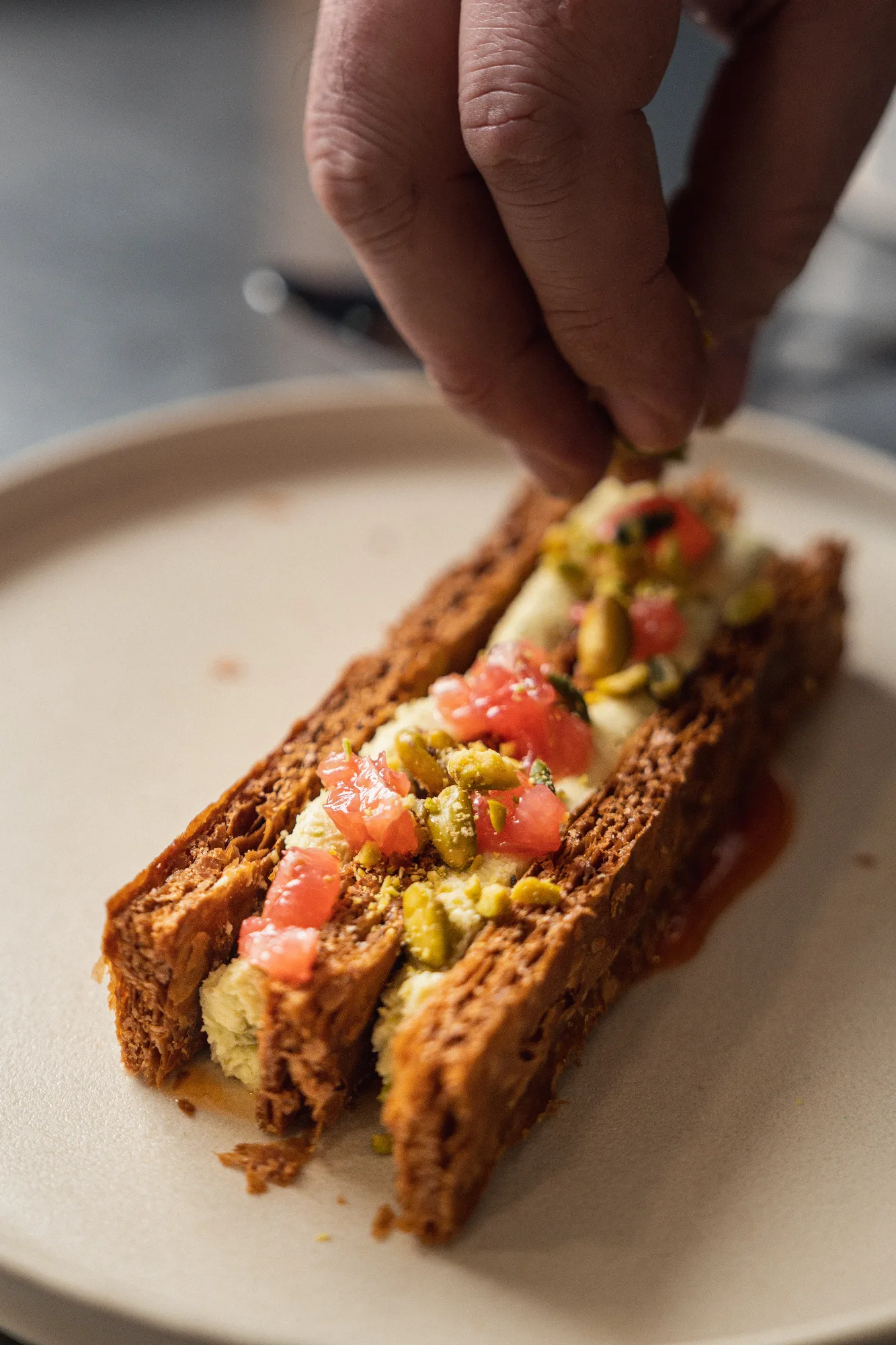 Une main dresse une tranche de pain toasté garnie de crème, pistaches concassées et dés de pamplemousse, sur assiette blanche.