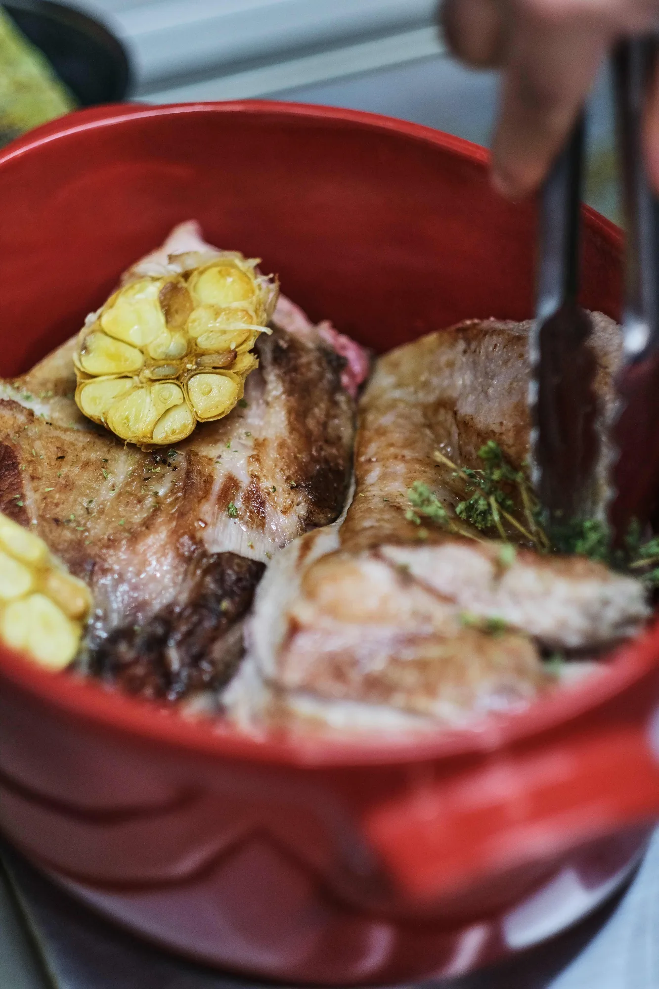 Une main saisit à la pince une tête d'ail rôtie au-dessus d'une pièce de viande grillée dans une casserole rouge, en cuisine bistronomique restaurant HABILE - vue 2