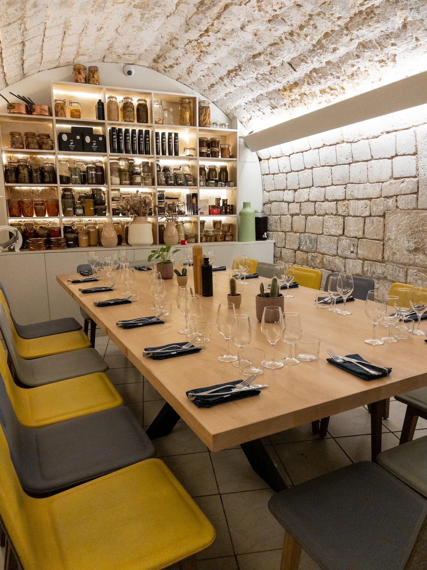 Salon privé du restaurant HABILE. : grande table en bois dressée sous voûte en pierre, chaises jaunes et grises, étagères d'épicerie fine éclairées en fond.