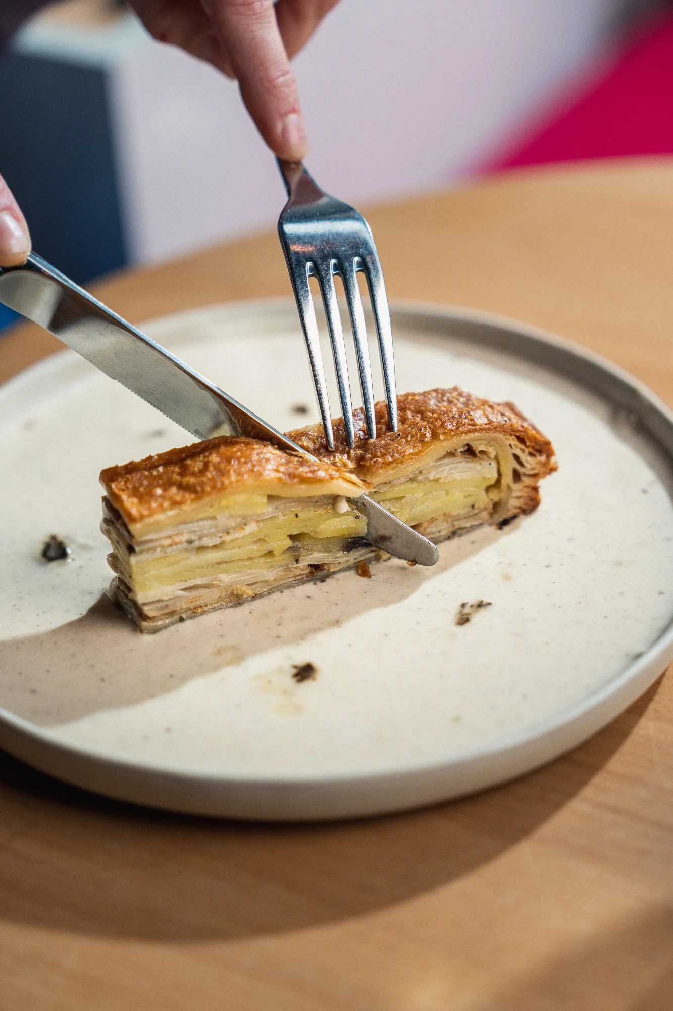 Tranche de feuilleté doré aux couches de pommes de terre et champignons, découpée au couteau et à la fourchette sur une assiette en grès, au restaurant HABILE.