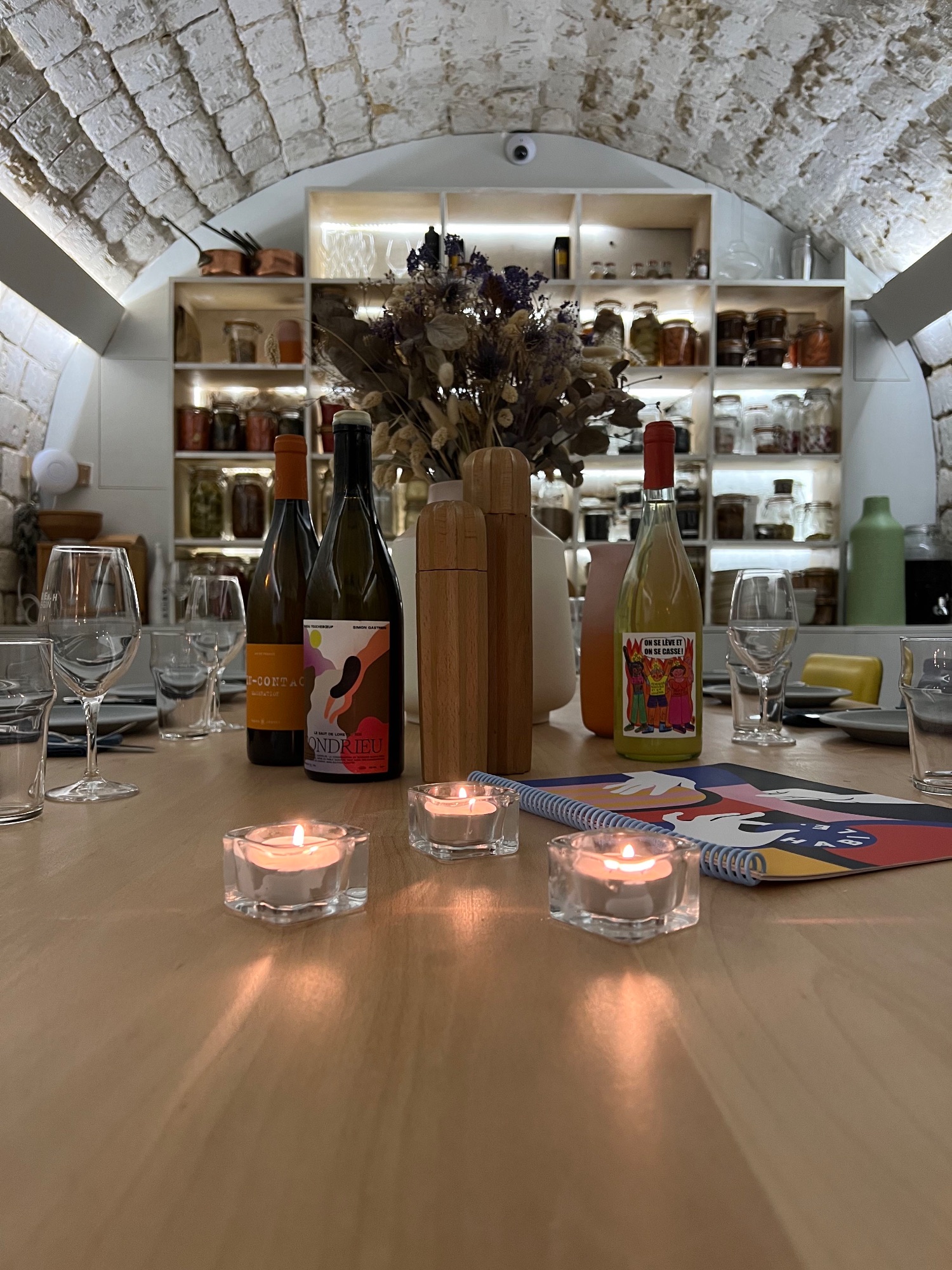 Salon privé d'HABILE. : table dressée aux bougies, trois bouteilles de vins vivants et étagères de conserves sous voûte en pierre.